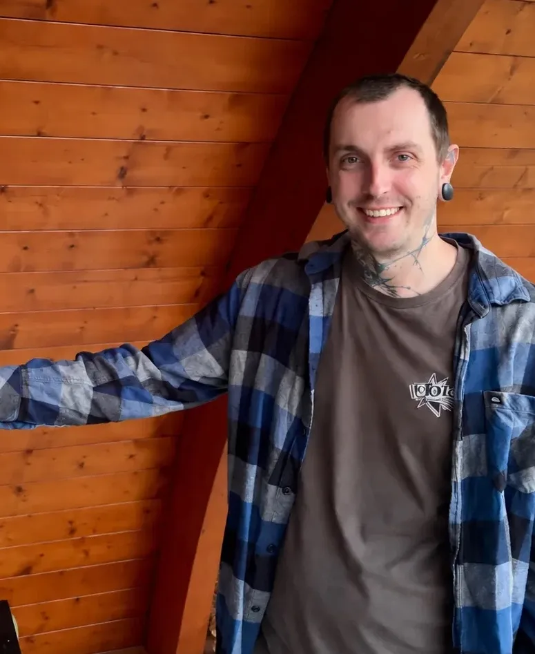 Bastien, souriant, en chemise à carreaux, sous un abri en bois